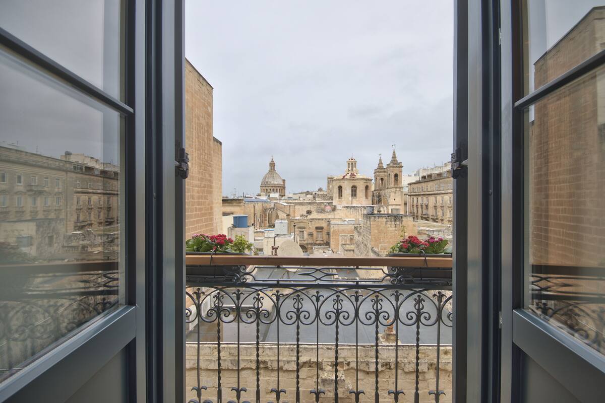 Une vue depuis un balcon orné de balustrades en fer forgé, donnant sur un paysage urbain historique avec d’imposants bâtiments à coupoles et à flèches.