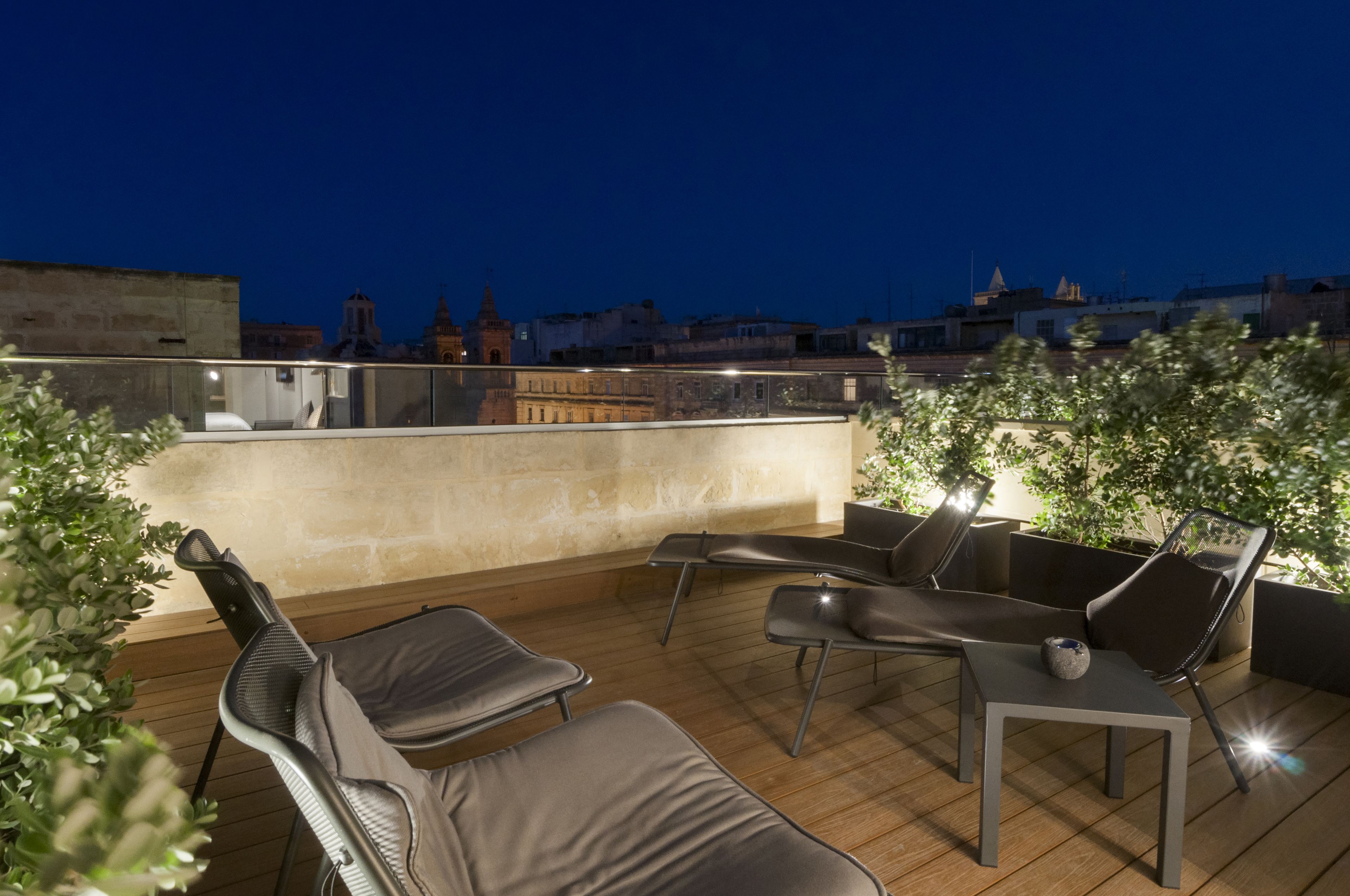 Terraza en la azotea con sillones de descanso y una mesita pequeña, con vistas a un paisaje urbano al anochecer.