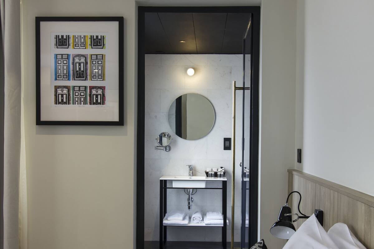 Une chambre d’hôtel avec un lit, un tableau encadré au mur, une salle de bain avec un lavabo et un miroir, et une porte donnant sur une autre pièce.