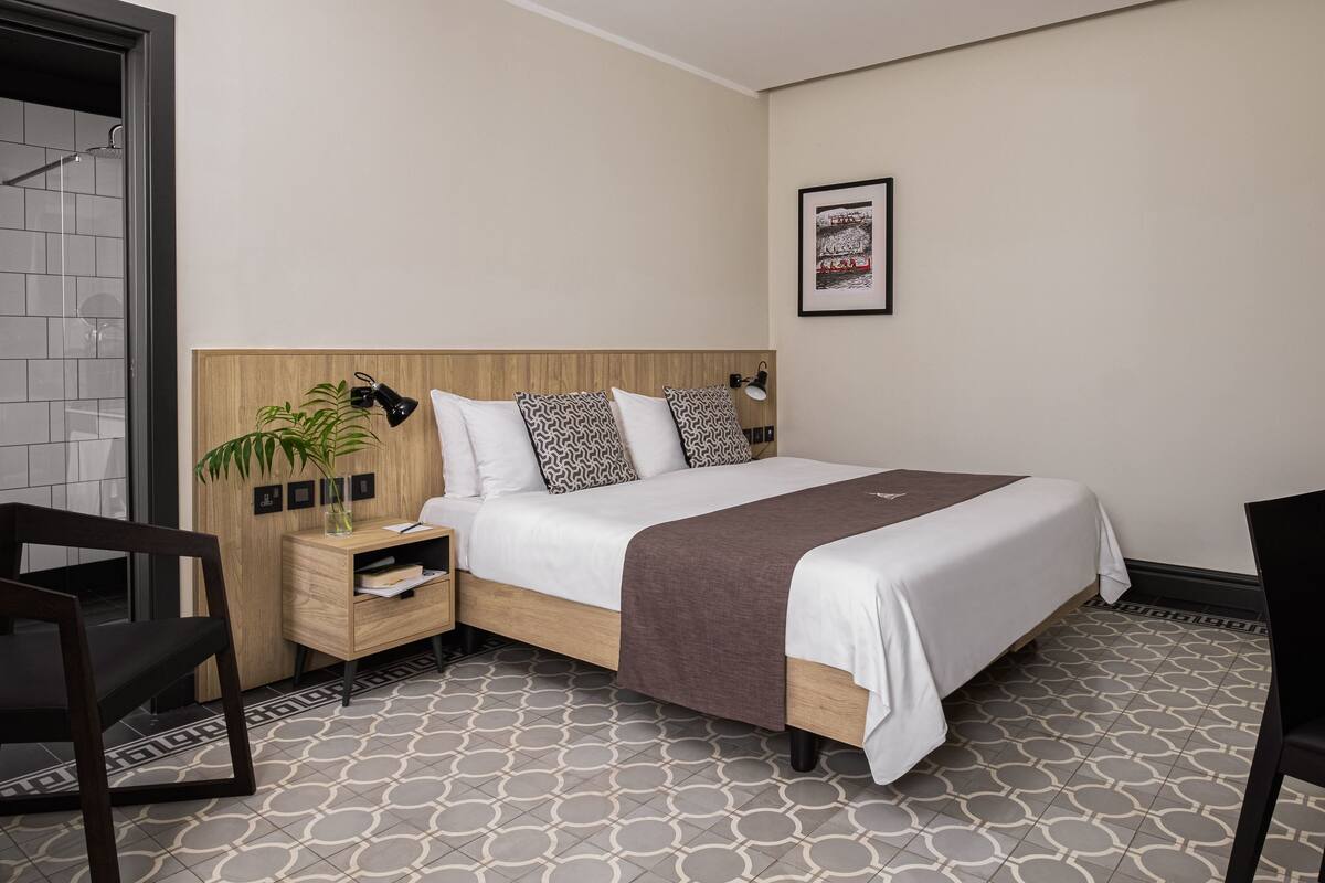 Une chambre d’hôtel moderne avec une tête de lit en bois, un grand lit agrémenté d’oreillers à motifs, une table de chevet avec une lampe et une petite plante.