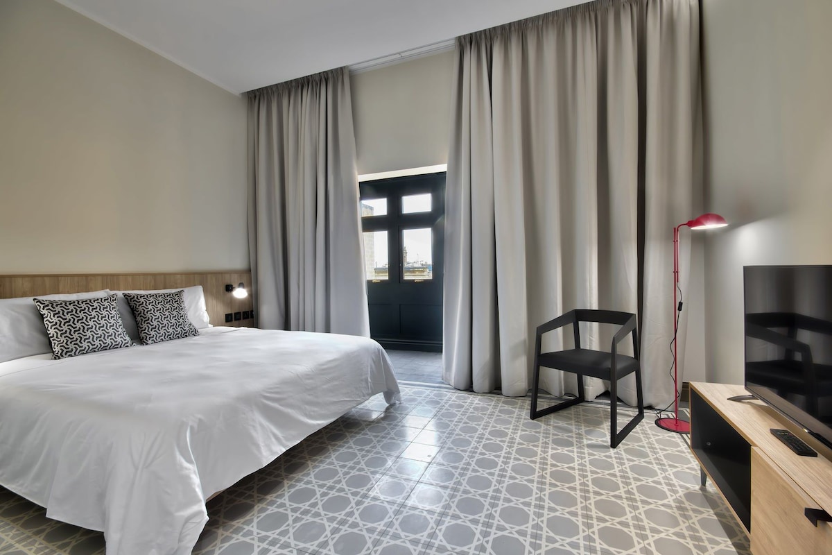 Une chambre d’hôtel avec un lit, une chaise, un bureau avec une télévision et une grande fenêtre avec des rideaux.