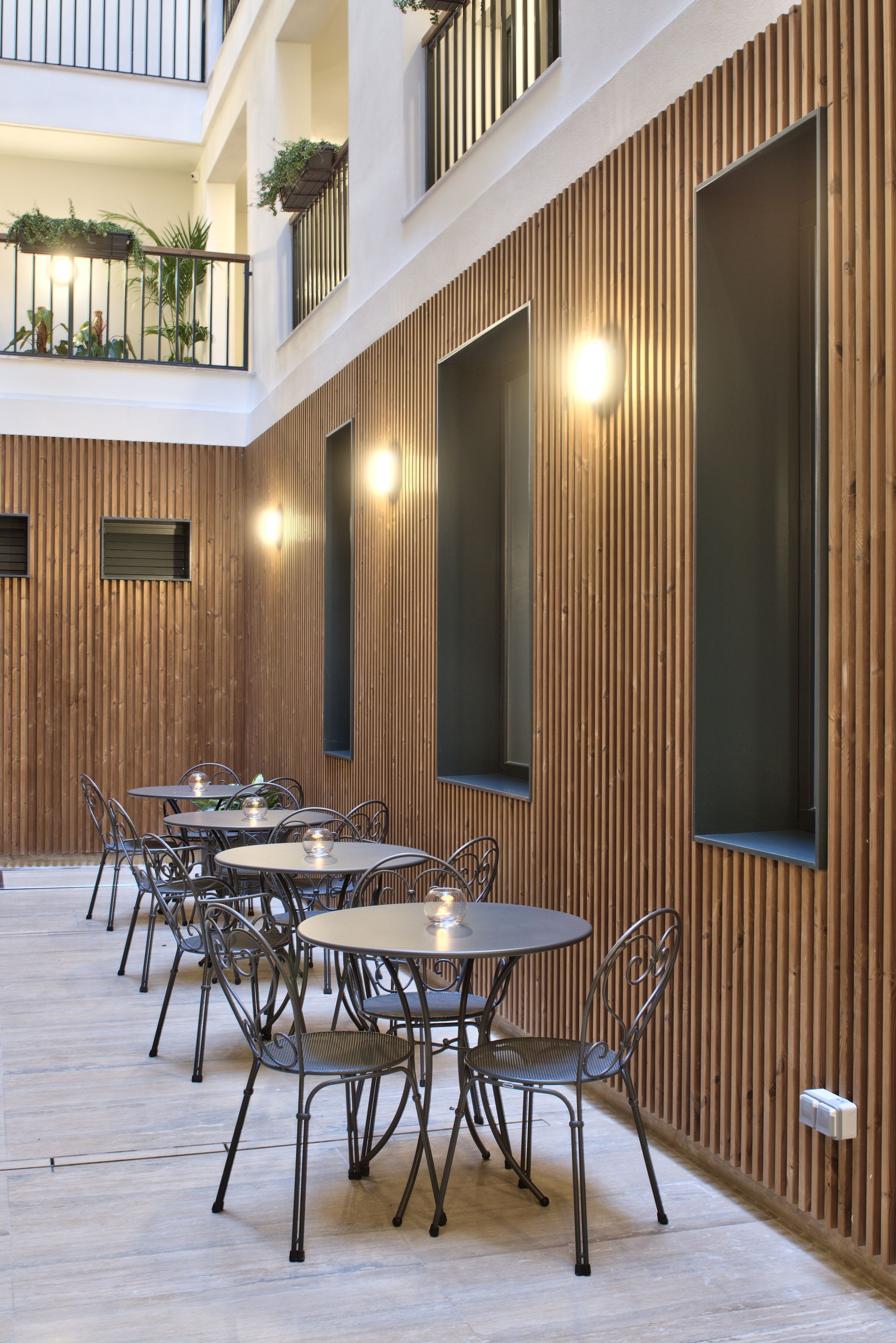A modern outdoor dining area with round tables and metal chairs, wooden paneling on the walls, and potted plants on the balconies.