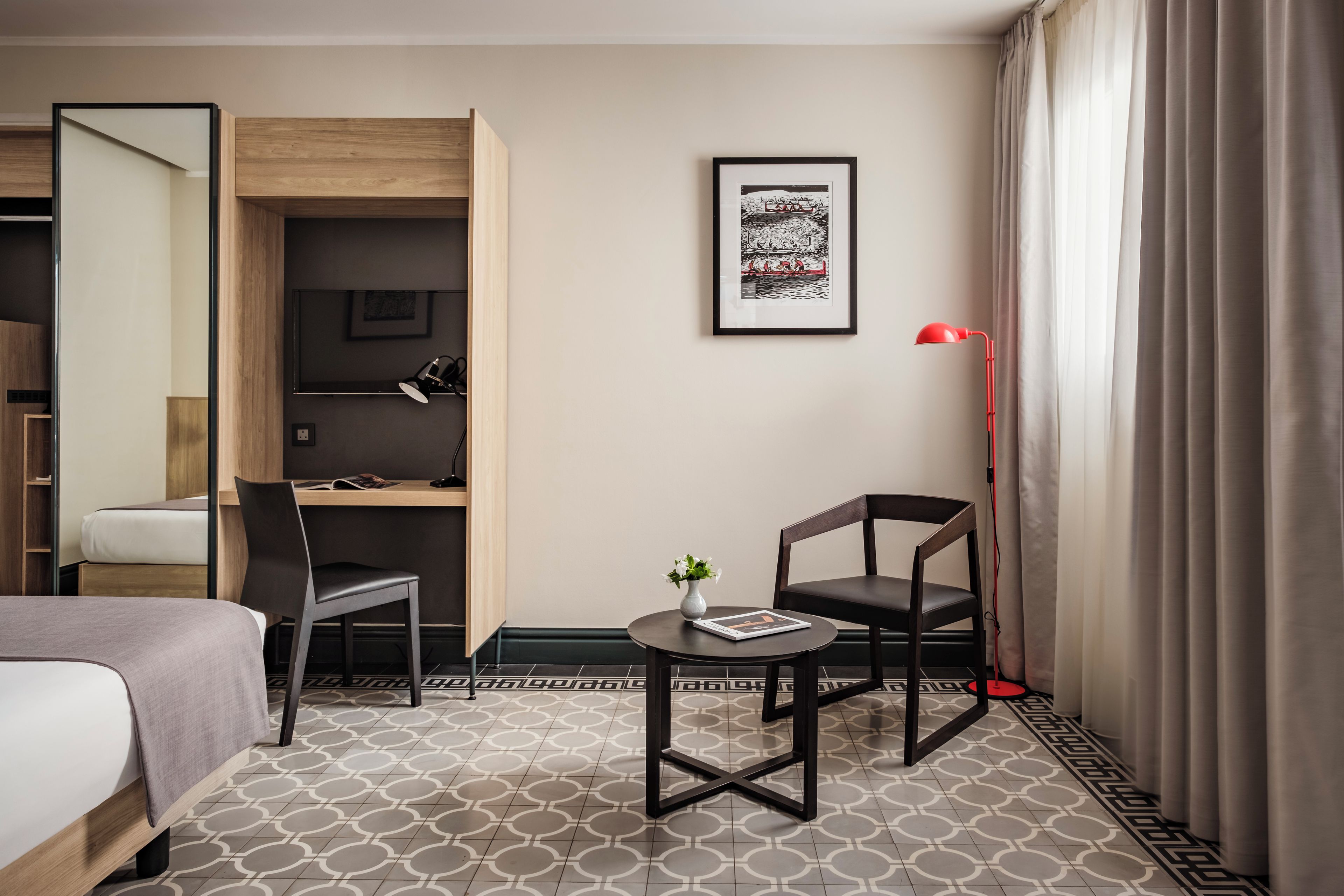 A hotel room with a bed, a desk, a chair, a small table with a vase, a red floor lamp, and a framed picture on the wall.
