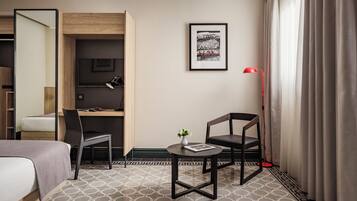 Habitación de hotel con cama, escritorio, silla, pequeña mesa con un florero, lámpara de pie roja y un cuadro en la pared.