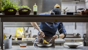 Un chef préparant des plats dans une cuisine professionnelle dotée de surfaces en acier inoxydable, d’une variété d’ustensiles de cuisine et de plantes en pot.