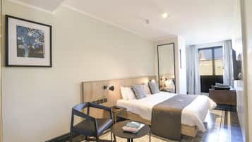 Habitación de hotel con cama, una silla, una mesita, un cuadro en la pared y un ventanal con cortinas.