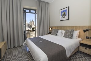 Habitación de hotel con una cama grande, cortinas grises, cabecera de madera, un cuadro enmarcado en la pared y vistas a un paisaje urbano a través de la ventana.