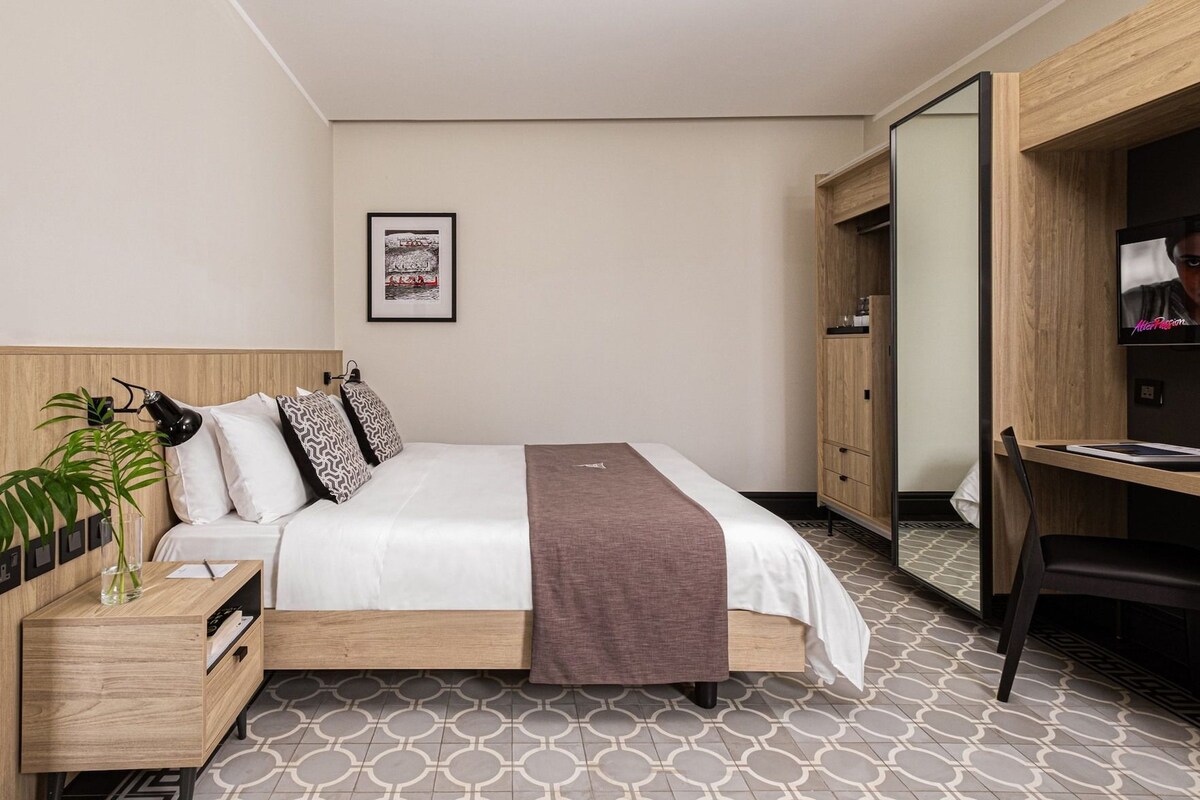 Une chambre d’hôtel comprenant un lit, une table de chevet, une lampe, un miroir, une armoire et un bureau avec une chaise.