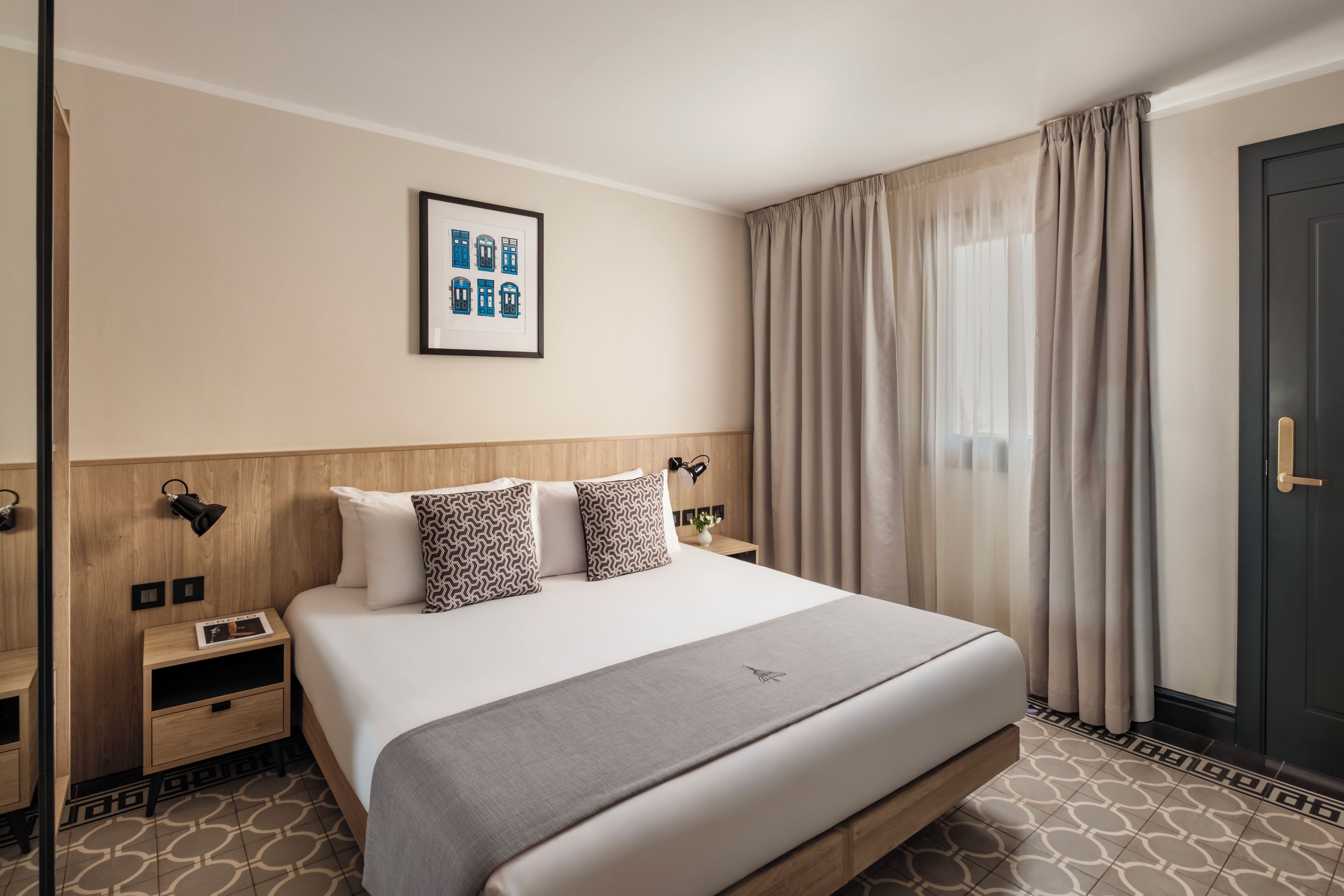 Une chambre d’hôtel avec un grand lit, une tête de lit, des tables de chevet, une petite table, un tableau encadré au mur et une fenêtre avec des rideaux.