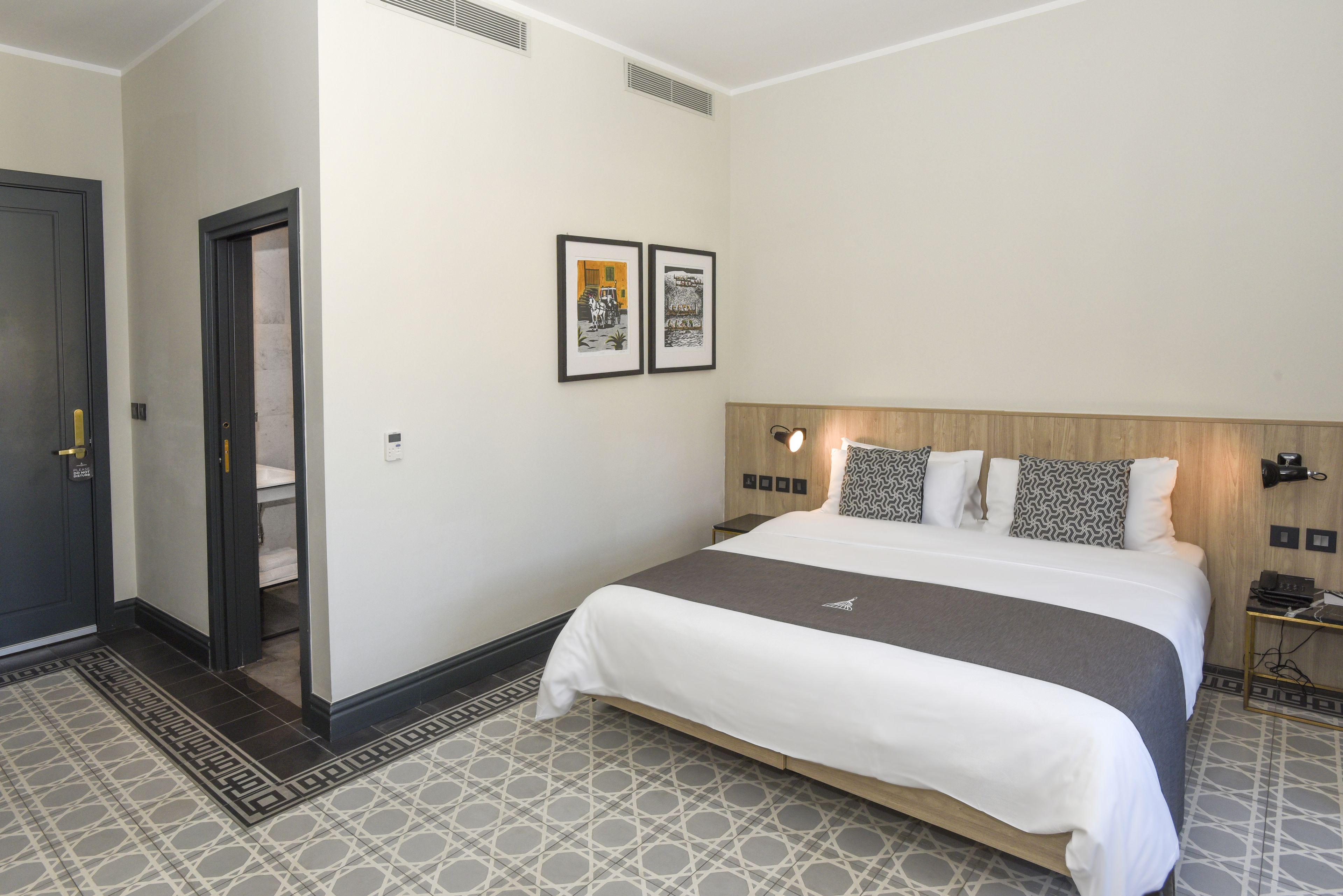 Hotellihuone, jossa on sänky, ovi ja kaksi kehystettyä kuvaa seinällä.
