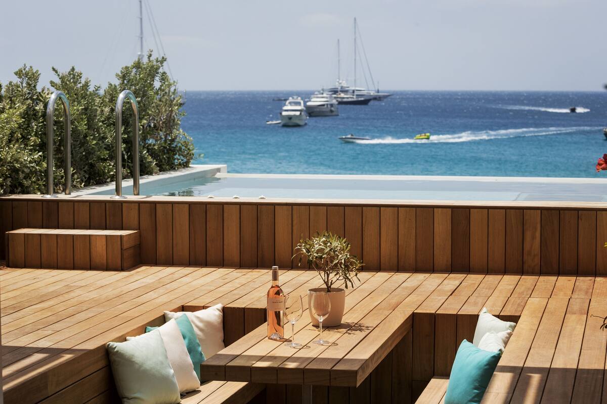 Una terraza de madera con asientos, una mesa con una botella y vasos, y una planta en maceta.