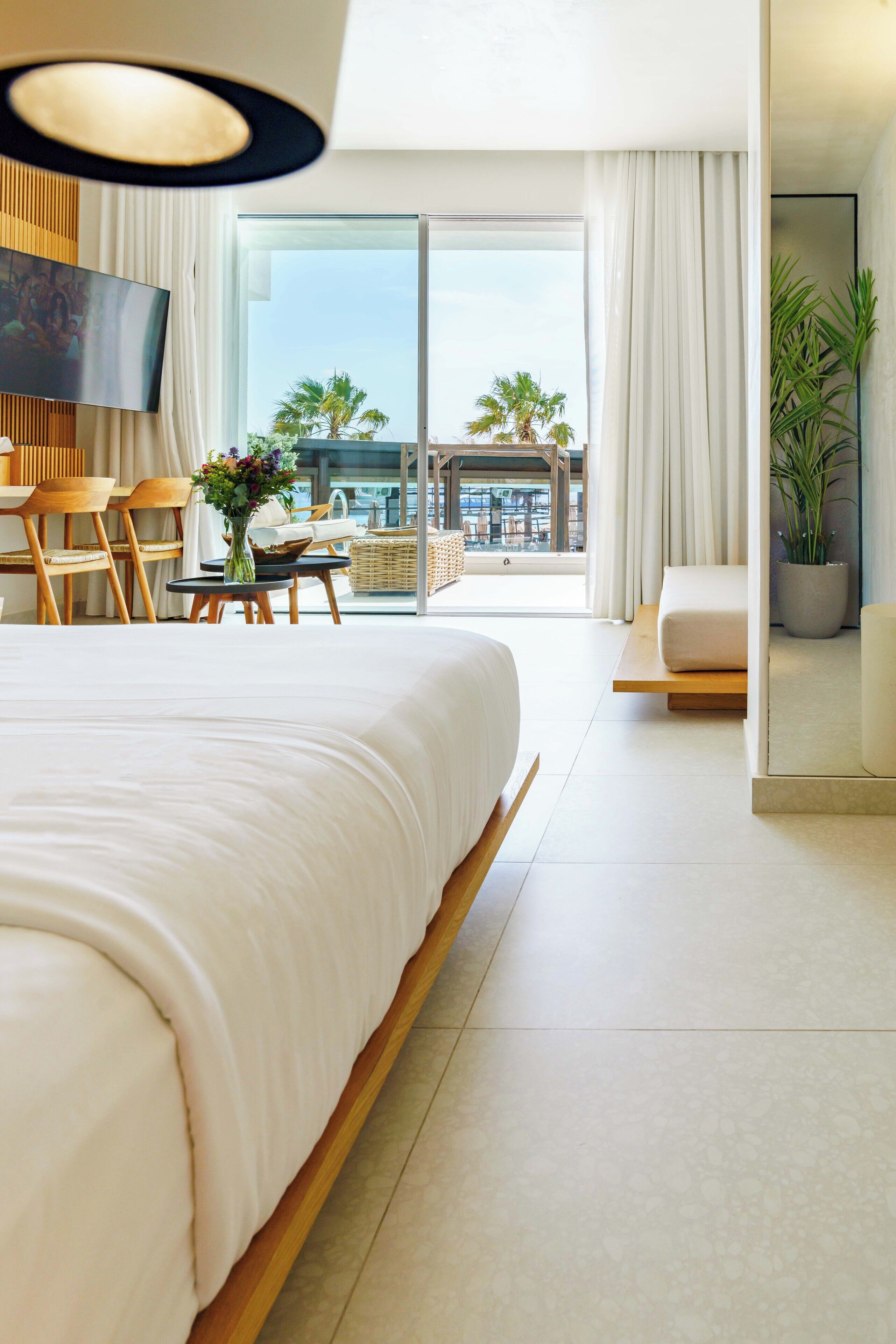 Een moderne hotelkamer met een groot bed, een flatscreen televisie, een zithoek met stoelen en een tafel, en een balkon met uitzicht op palmbomen en gebouwen.