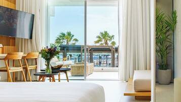 Een moderne hotelkamer met een groot bed, een flatscreen televisie, een zithoek met stoelen en een tafel, en een balkon met uitzicht op palmbomen en gebouwen.