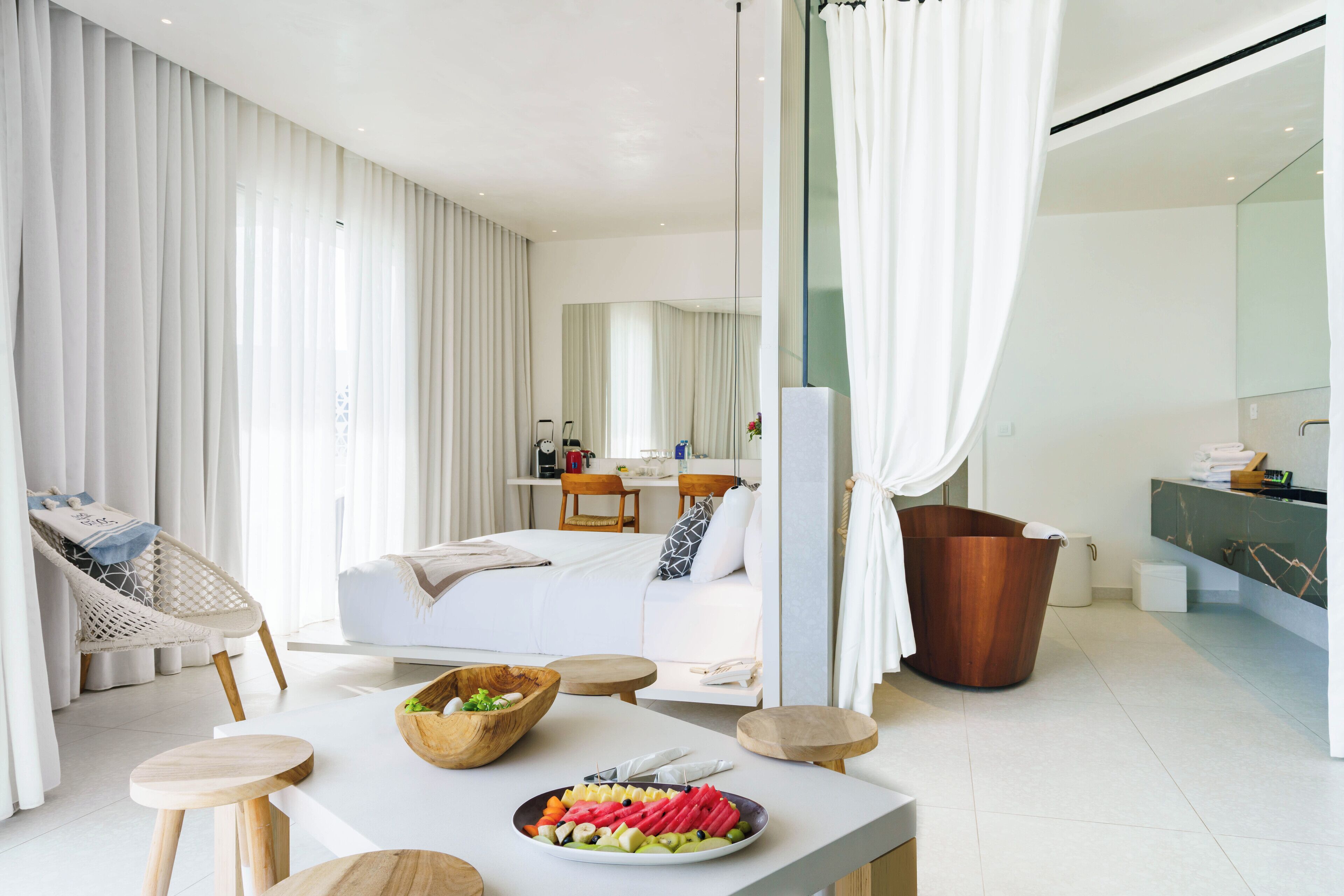 A modern hotel room with a dining area, a sitting area with a sofa and chairs, a large mirror, and a wooden bucket.