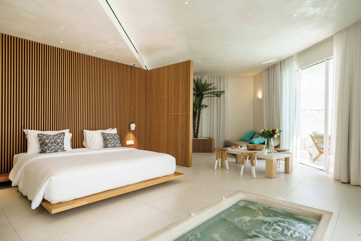 Habitación de hotel moderna con una cama grande, una mesita con sillas, una planta y vistas al exterior.