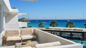 A balcony with a white sofa and cushions, a small table with a tray, and a view of the sea with boats and palm trees. - Mykonos Dove Beachfront Hotel (Mykonos)