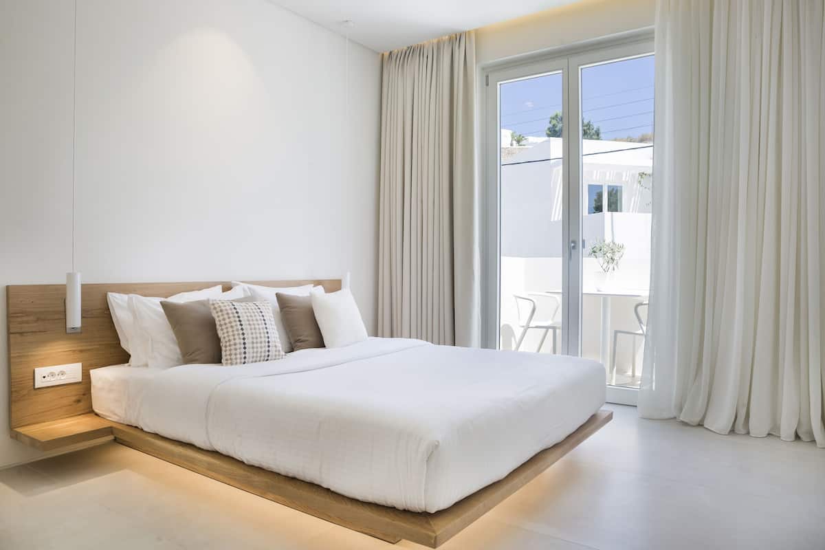 Un dormitorio moderno con cabecera de madera, cama grande con ropa de cama blanca, mesita de noche y vistas al exterior a través de una puerta corrediza de cristal.