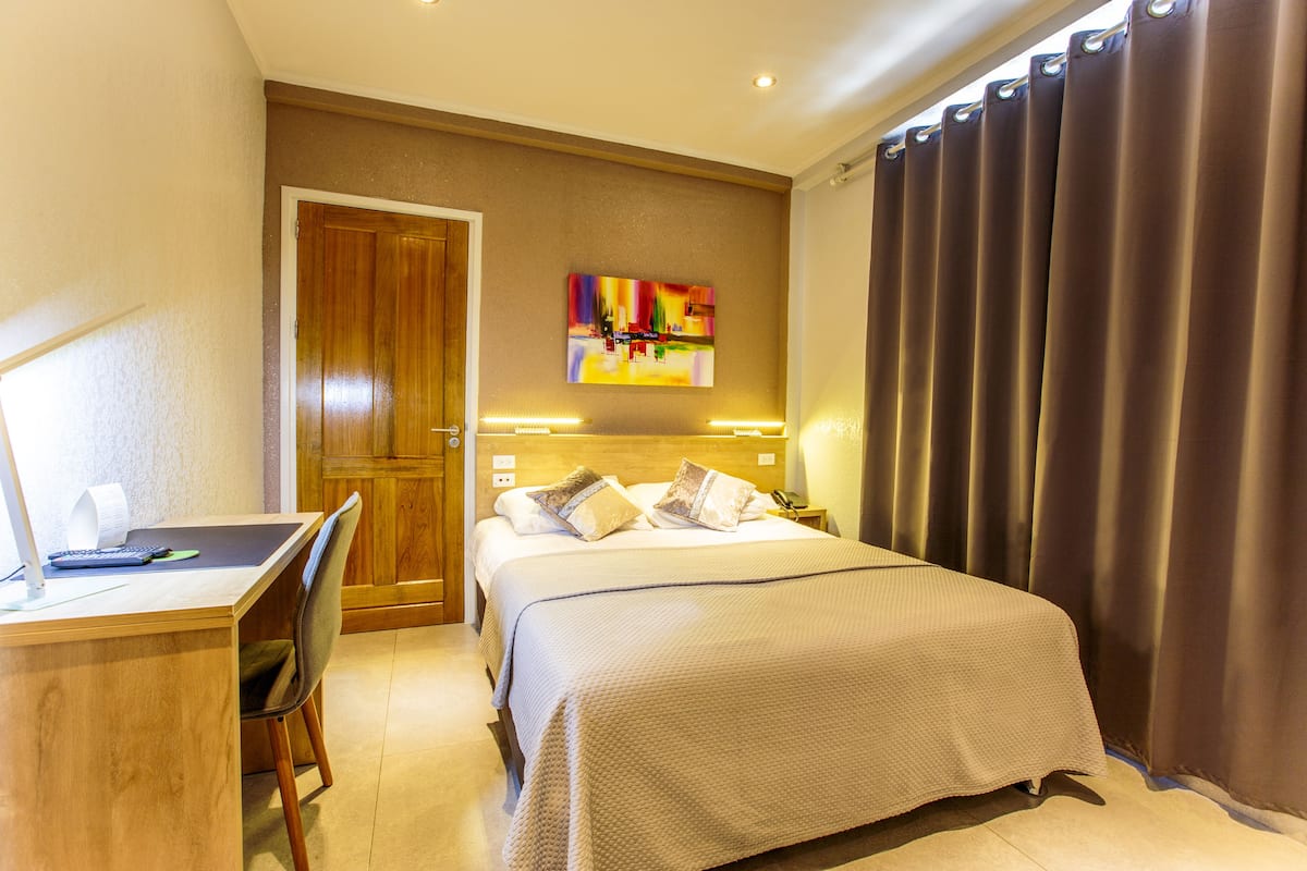 A hotel room with a bed, desk, chair, and artwork on the wall.