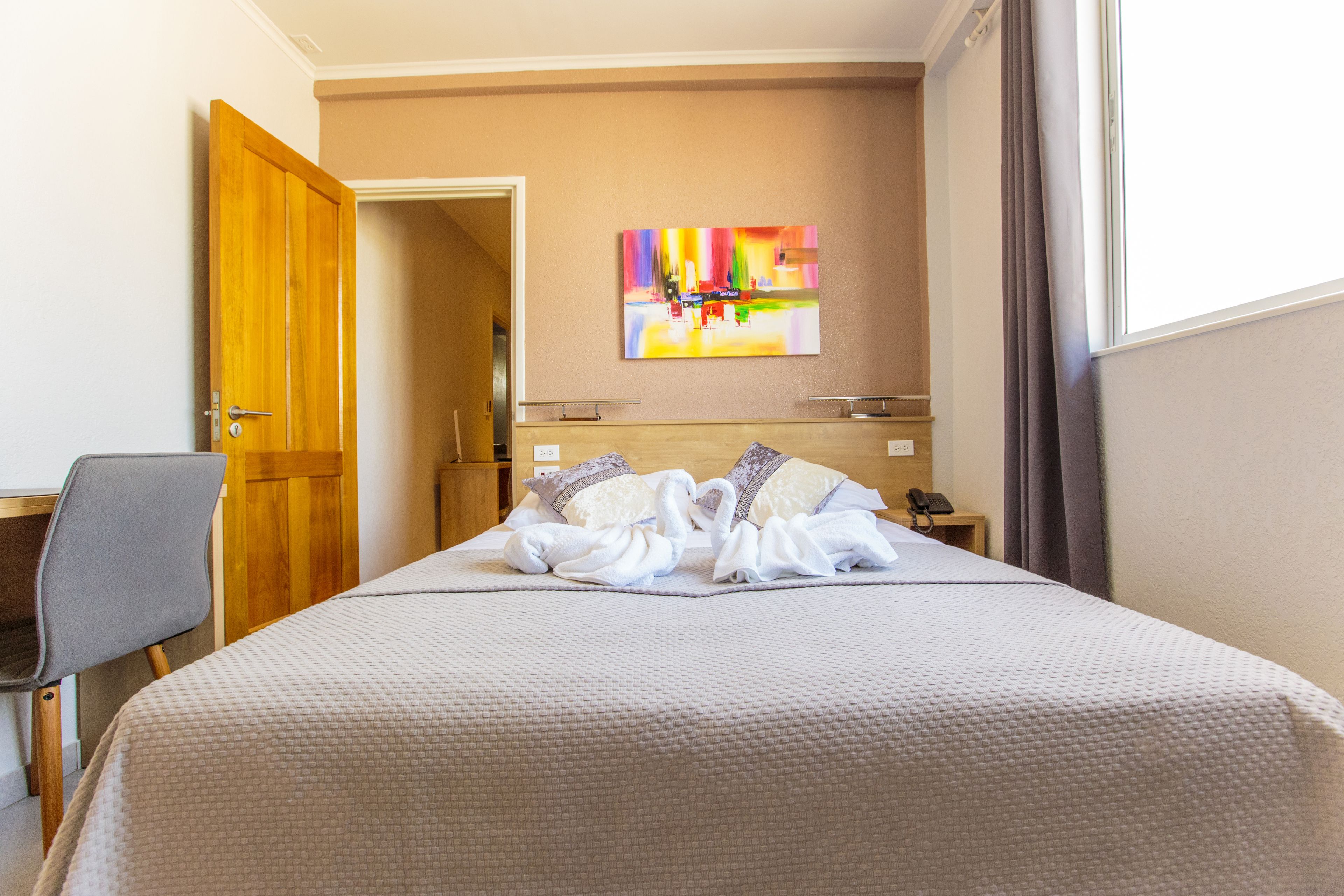 A hotel room with a bed, a desk, a chair, a painting on the wall, and a window with curtains.