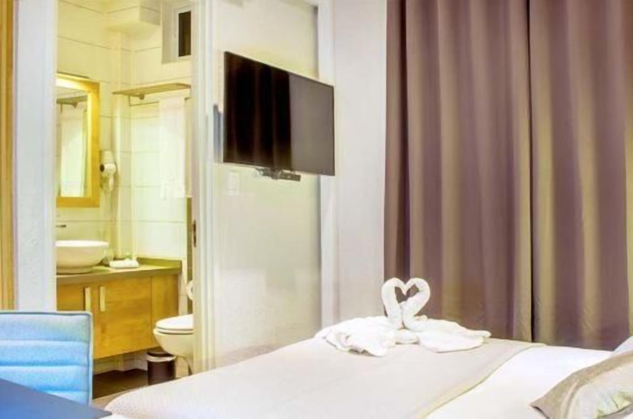 Una camera d'albergo con un letto, una TV a muro, un bagno con lavandino e specchio, e una finestra con tende.