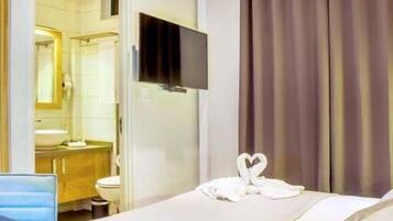 Una camera d'albergo con un letto, una TV a muro, un bagno con lavandino e specchio, e una finestra con tende.