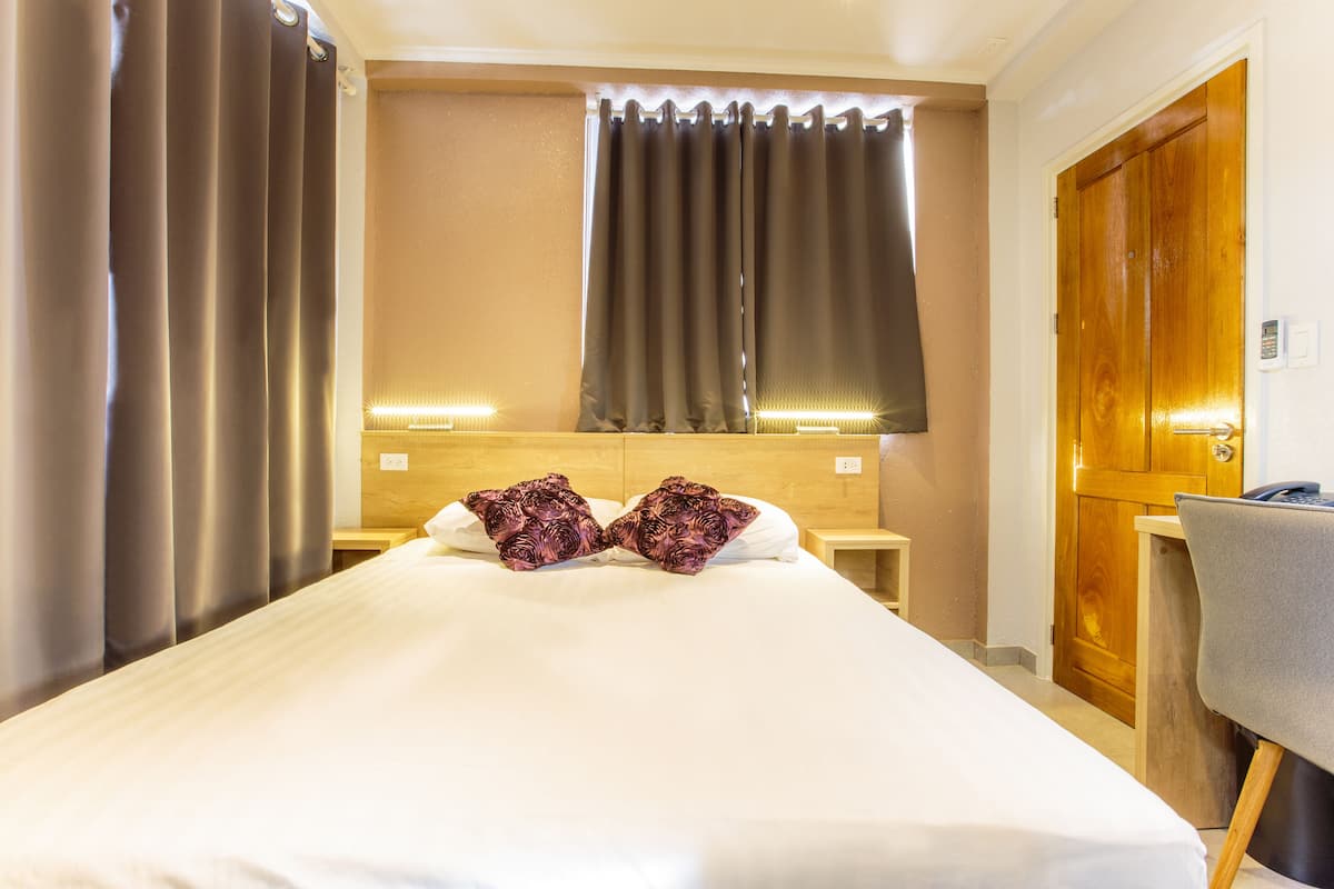 A hotel room with a large bed, two decorative pillows, a wooden headboard, a desk with a chair, and a wooden door.