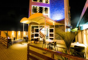 An outdoor dining area with tables and chairs under a canopy, a staircase, and a building with large windows and a brick facade.