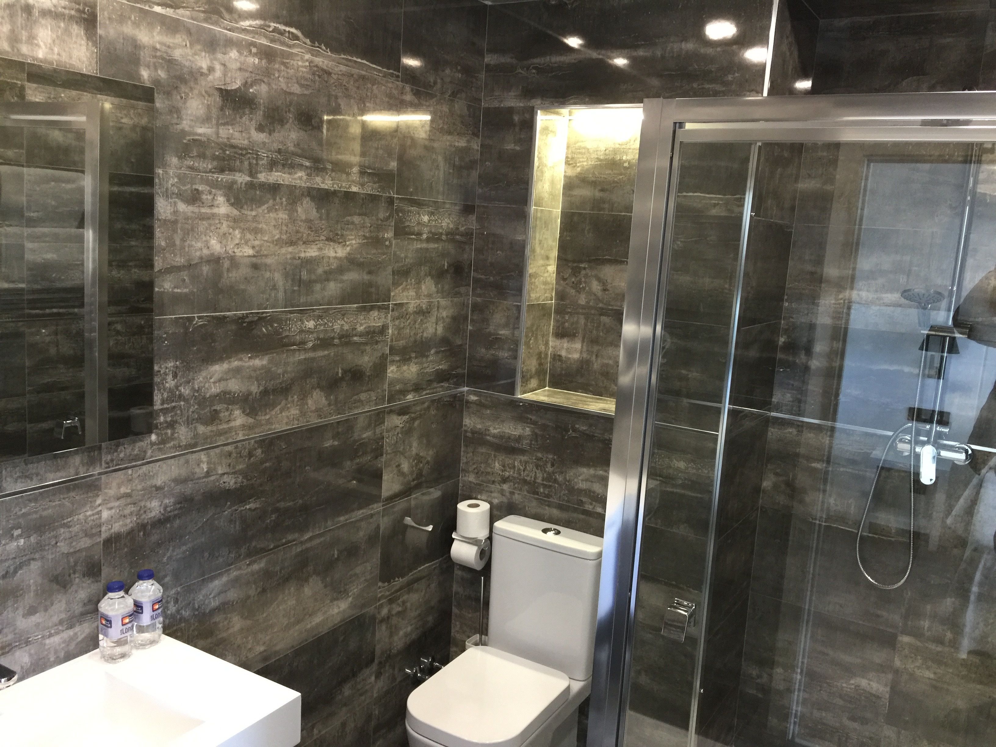A modern bathroom with dark marble tiles, a glass shower enclosure, a white toilet, and a sink with bottled water.