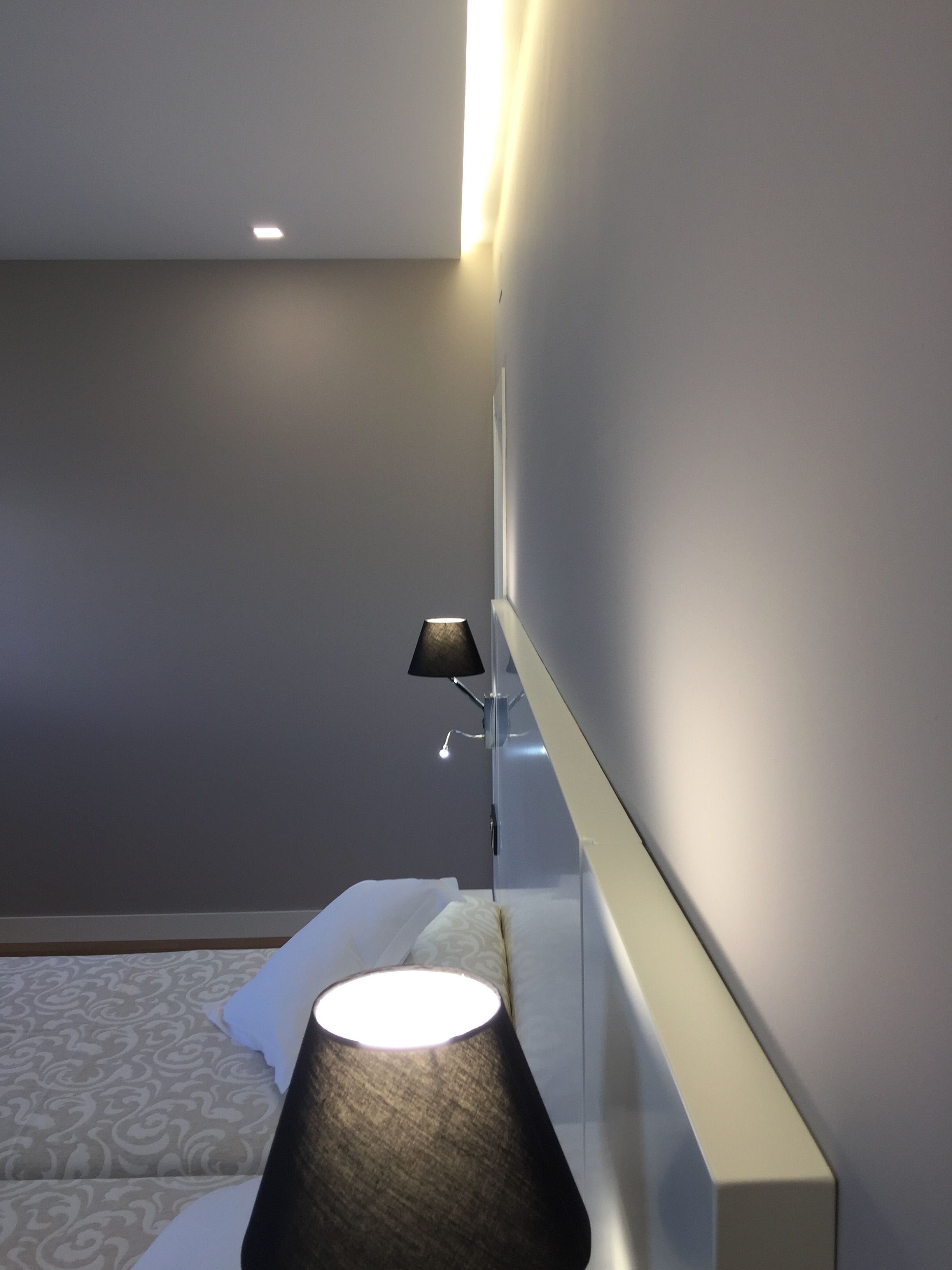 A modern hotel room with a bed, a bedside table with a lamp, and a wall-mounted light fixture.