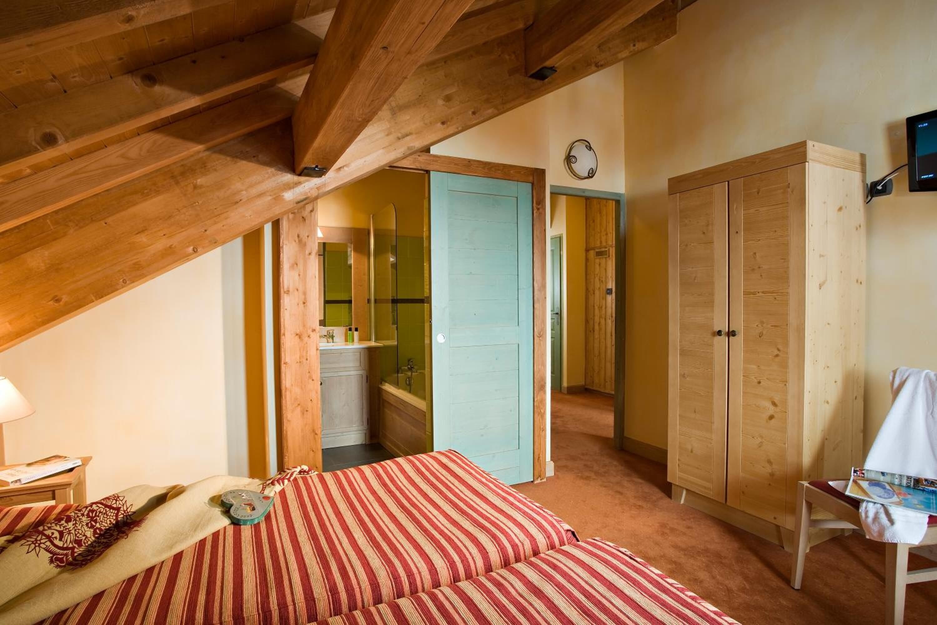 Ein gemütliches Zimmer mit schrägen Wänden, einem Bett mit gestreifter Bettwäsche, einem Holzschrank und einem kleinen Tisch mit einer Zeitschrift.
