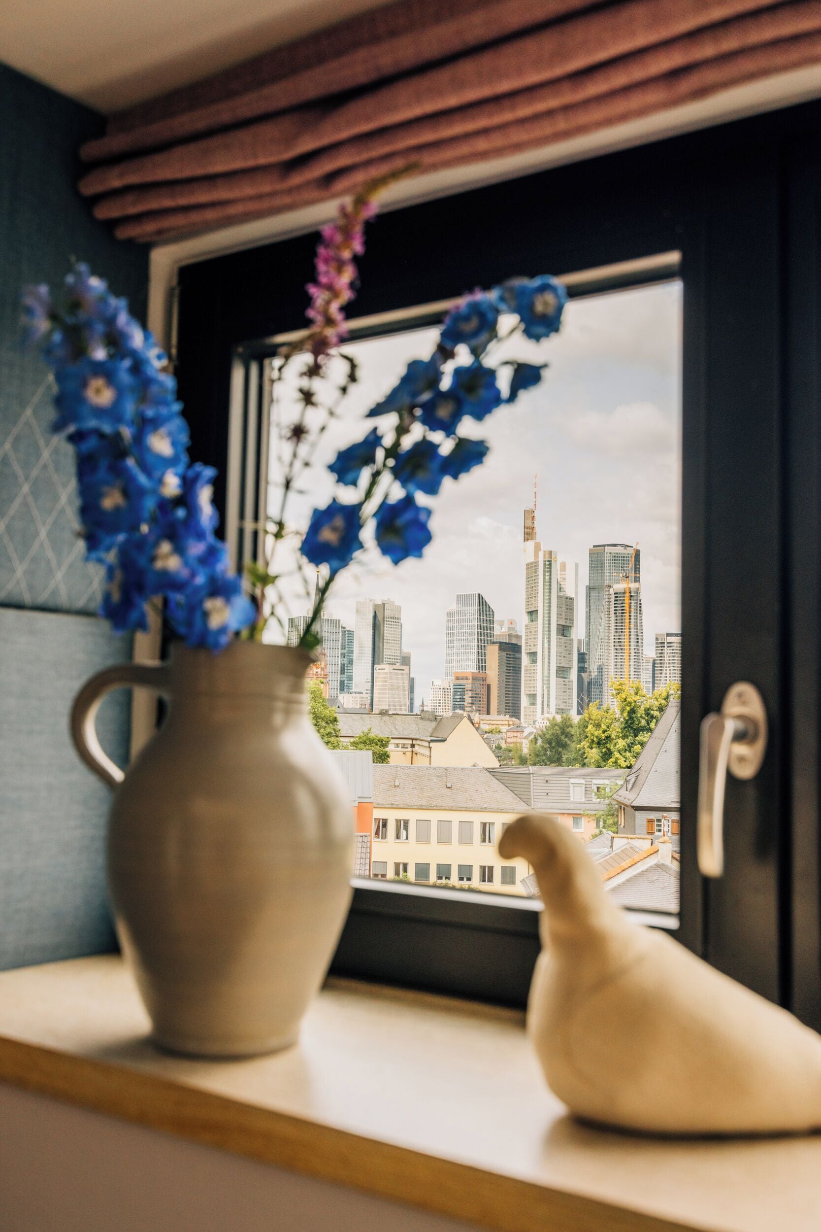 Een vaas met bloemen op een vensterbank, een vogelbeeldje en een stadsgezicht door een raam.