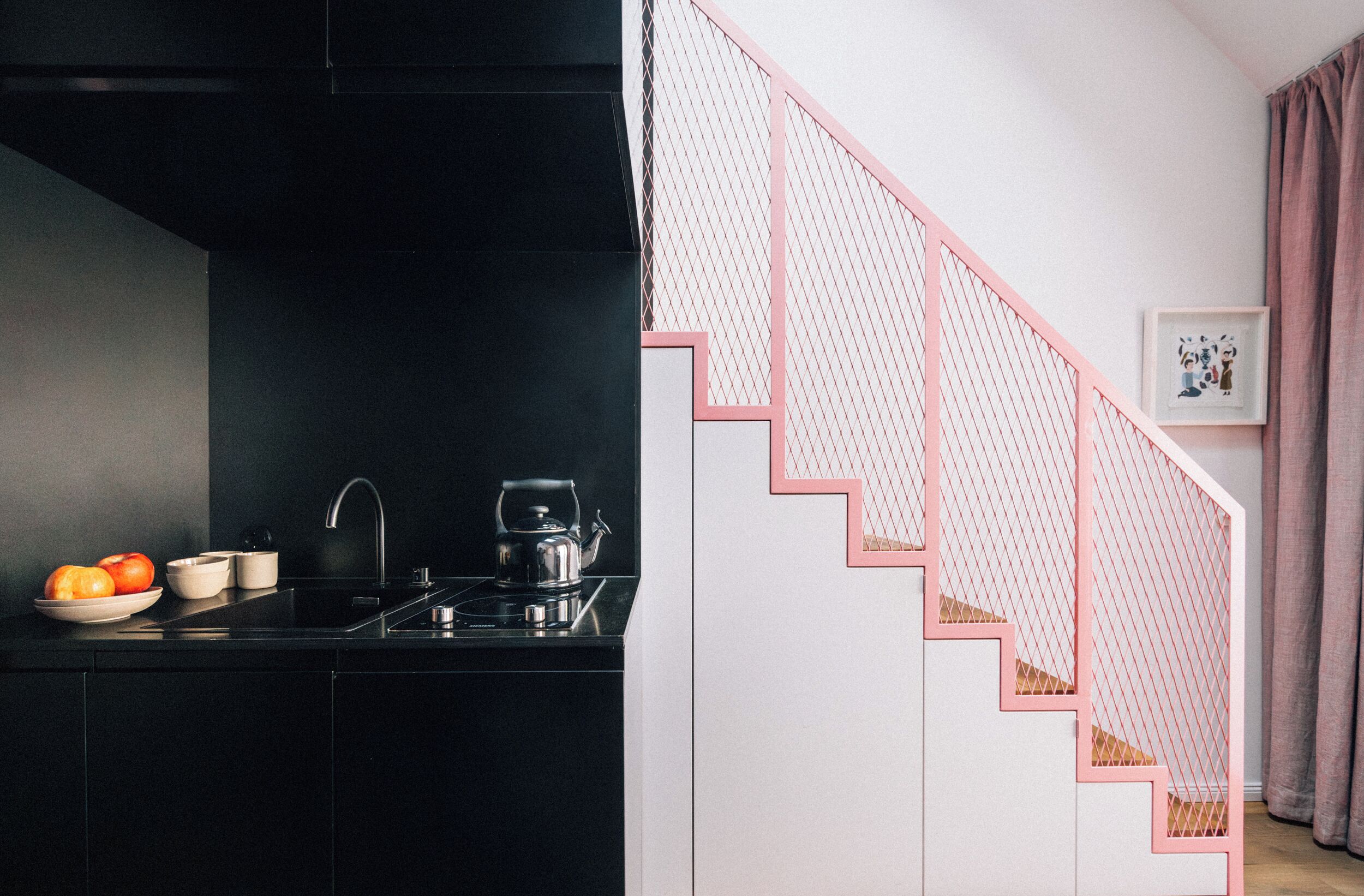 Una cocina con encimera negra, un lavamanos y una tetera. Se ve una escalera con barandillas rosas que conduce a otro nivel.