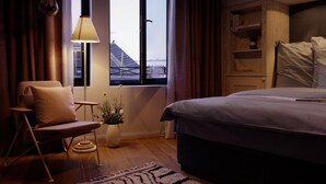 Una habitación de hotel moderna con cama, una silla, una lámpara, una ventana con cortinas y vistas a edificios.