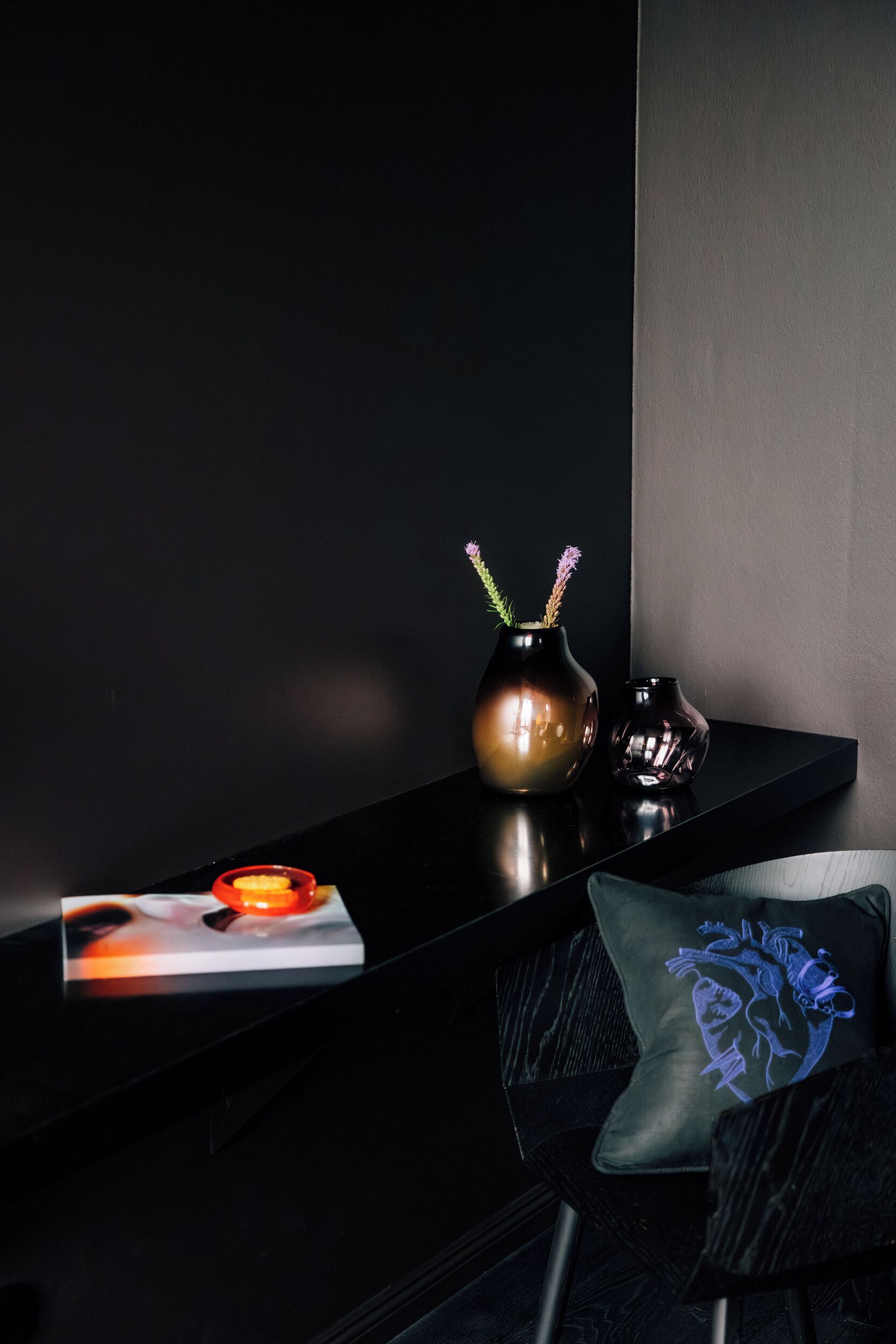 A modern, dark-themed room with a sleek black console table holding a vase with flowers, a small decorative item, and a pillow with a blue design.