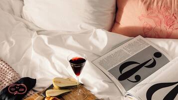 A neatly made bed with white linens, a magazine, a tray with croissants, apples, and a glass of red wine.