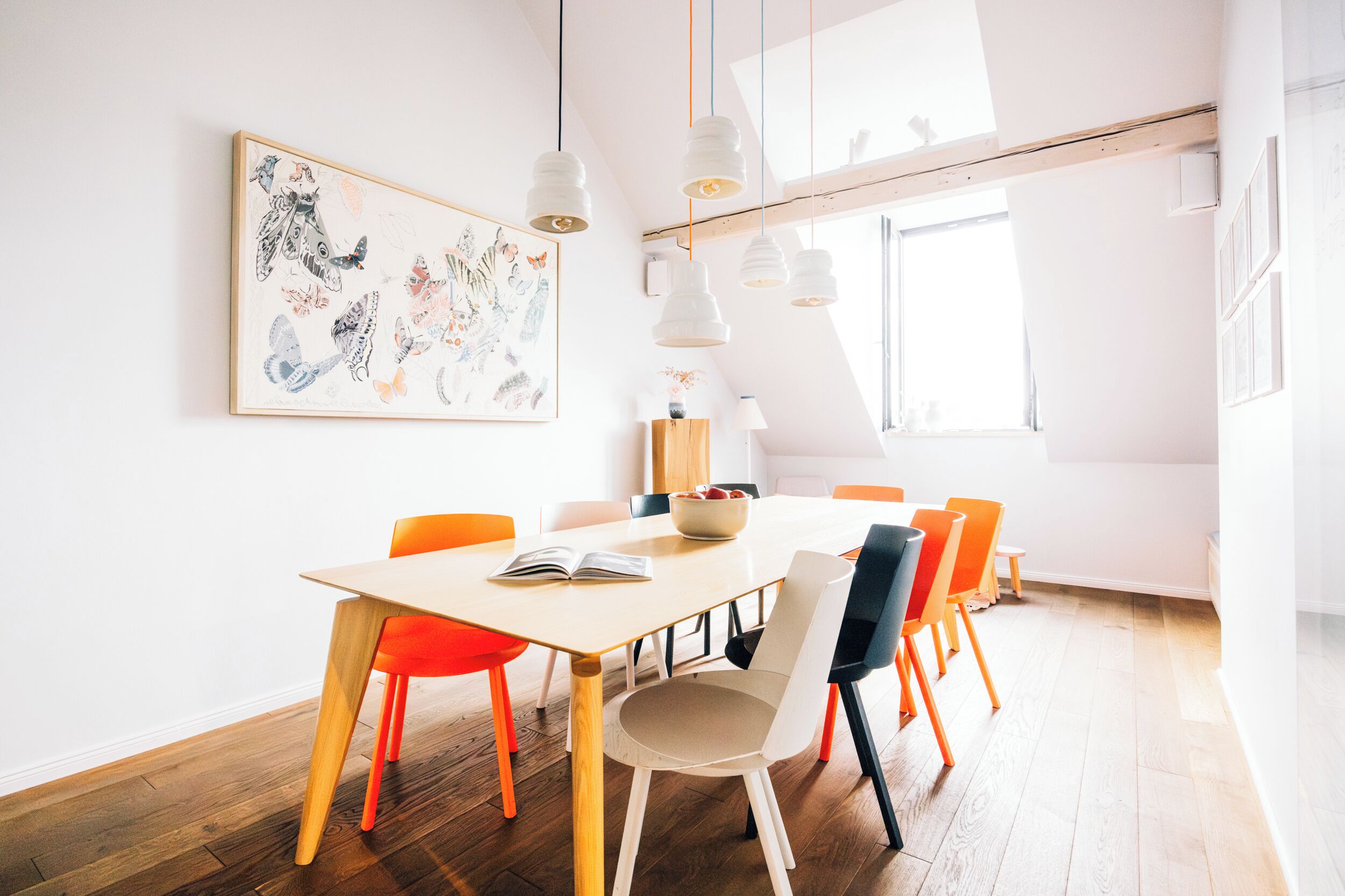 A modern dining area with a wooden table, orange and white chairs, and a large framed artwork on the wall.