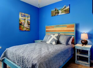 A bedroom with a blue wall, a bed with a grey quilt, two pillows, a wooden headboard, a nightstand with a lamp, and two paintings. - Bonzai Bay SP1 2 8 9 (Port Aransas)