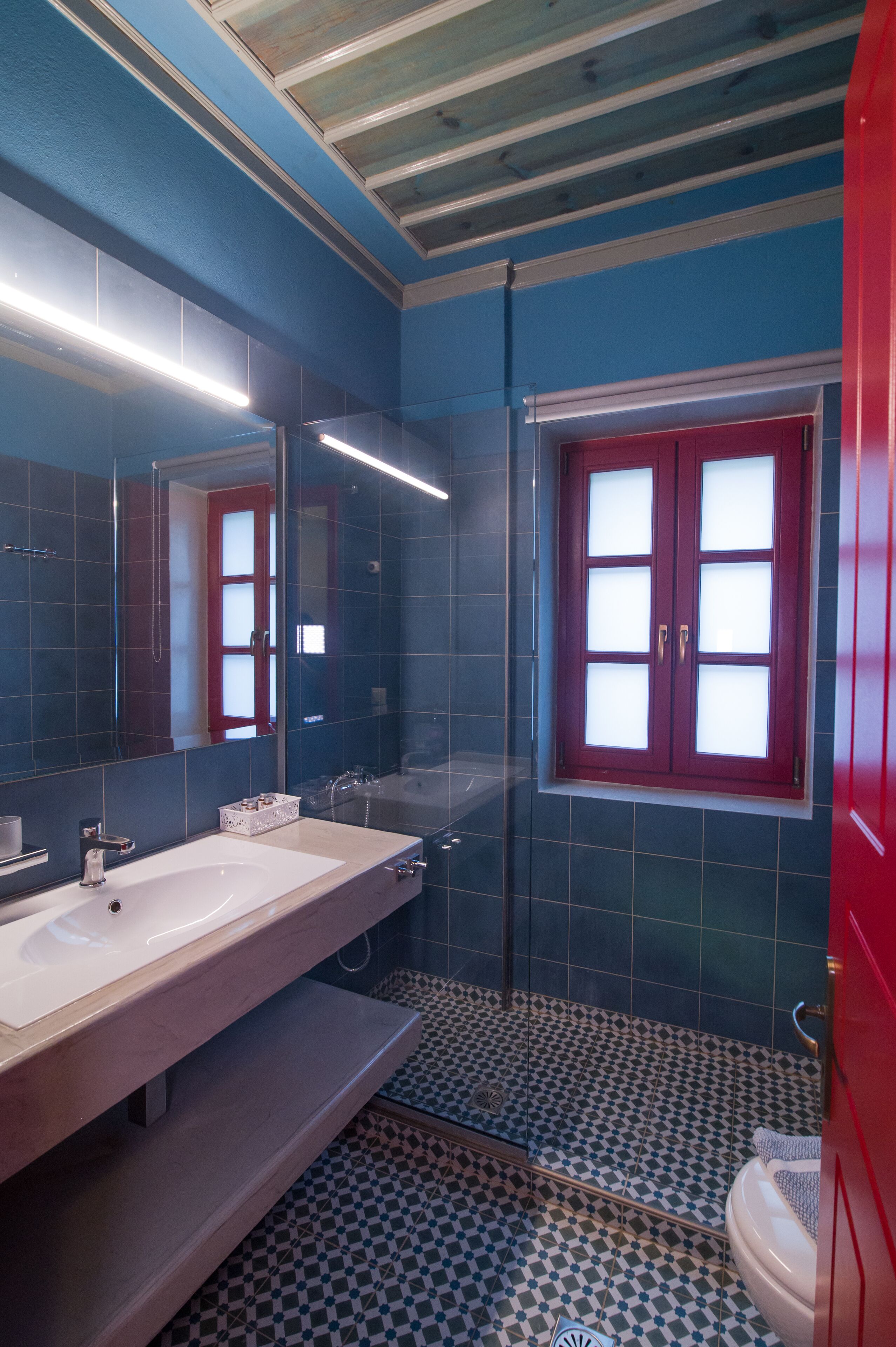 A bathroom with a large mirror, a white sink, a red-framed window, and a patterned floor.