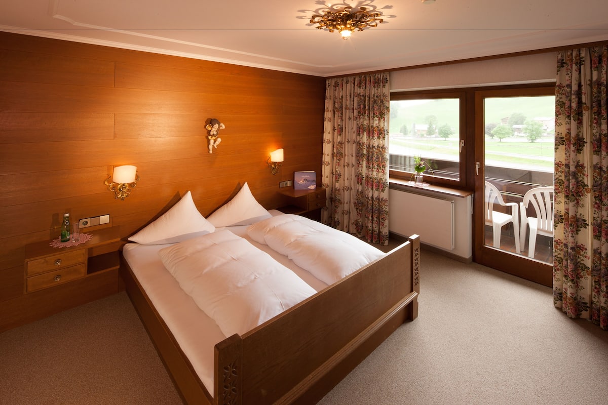 A hotel room with a wooden headboard, a large bed with white linens, two wall-mounted lamps, a small bedside table with a bottle and a glass, and a window with floral curtains.
