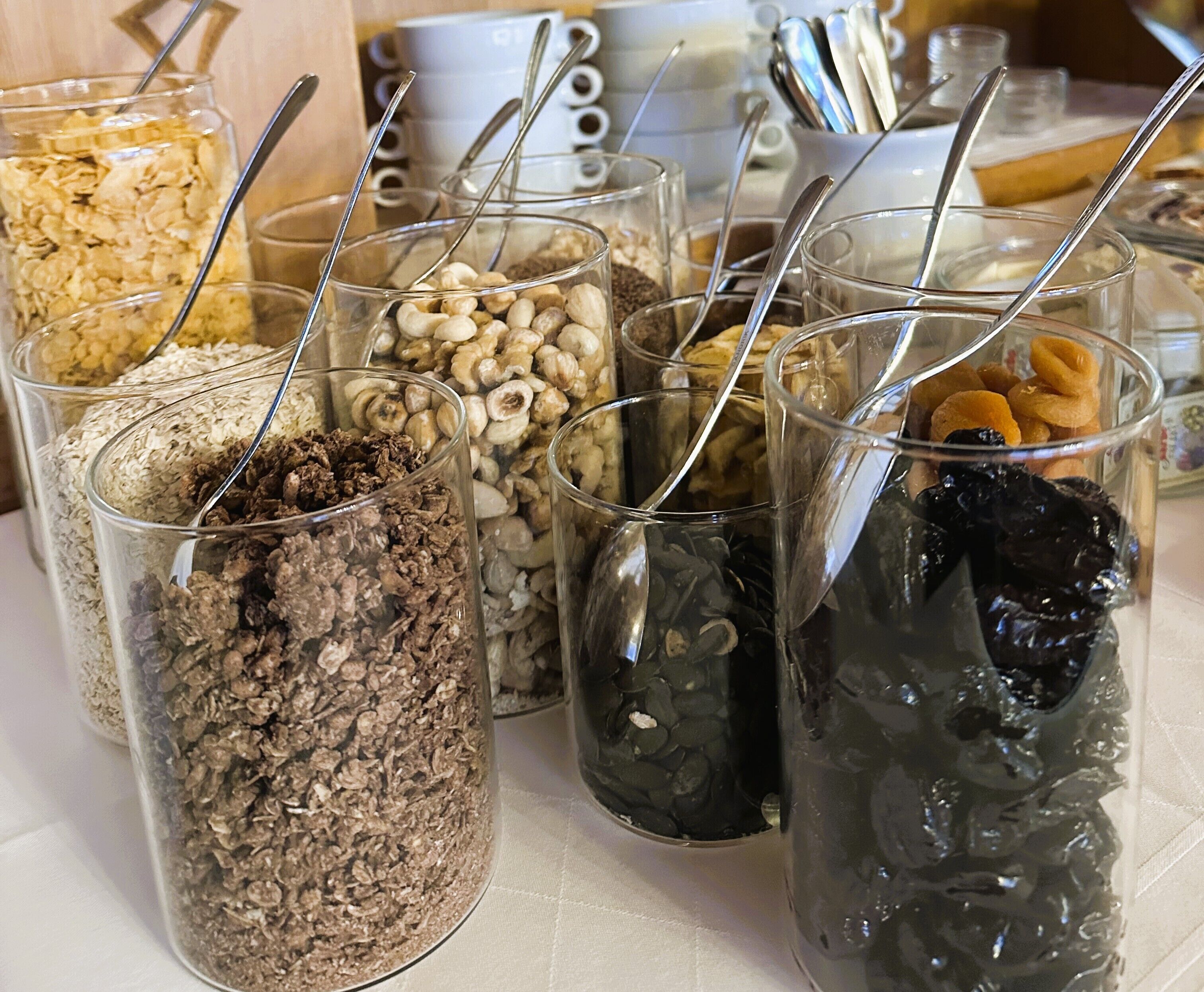 A variety of food items in glass containers with spoons, including cereals, nuts, and dried fruits.
