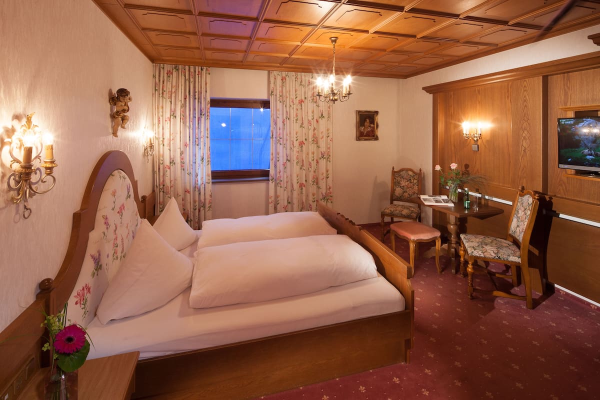 A hotel room with a double bed, wooden headboard, floral curtains, a wooden desk with a chair, a television, and a chandelier.