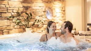 A couple in a hot tub with a stone wall and plants in the background. - Hotel Elisabeth (Schoppernau)
