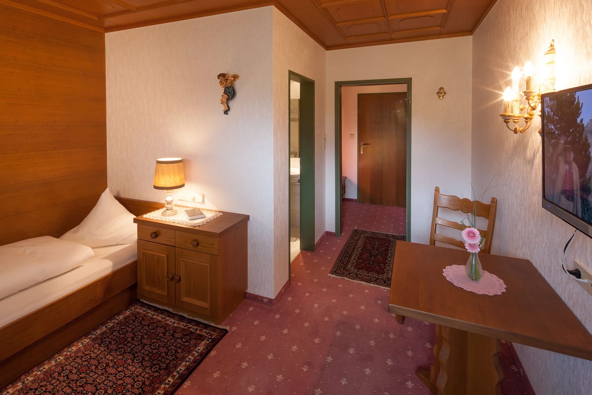 A room with a bed, a wooden nightstand with a lamp, a small table with a vase, a chair, and a television on the wall.