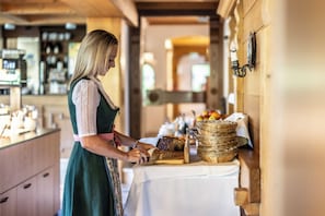 A woman slicing bread at a counter with baskets of fruits and a coffee machine in the background. - Hotel Elisabeth (Schoppernau)