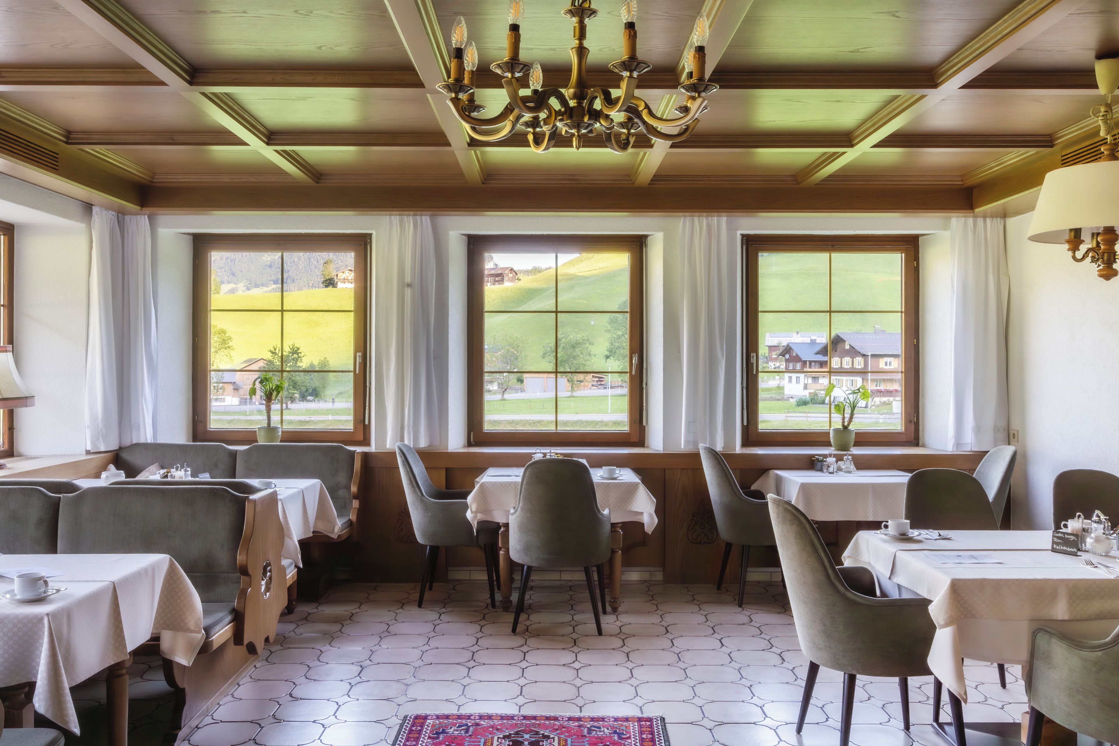 A dining area with tables and chairs, large windows with curtains, a chandelier, and a view of a green landscape.