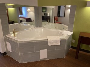 A corner bathroom with a large bathtub, a mirror, and a wooden floor.