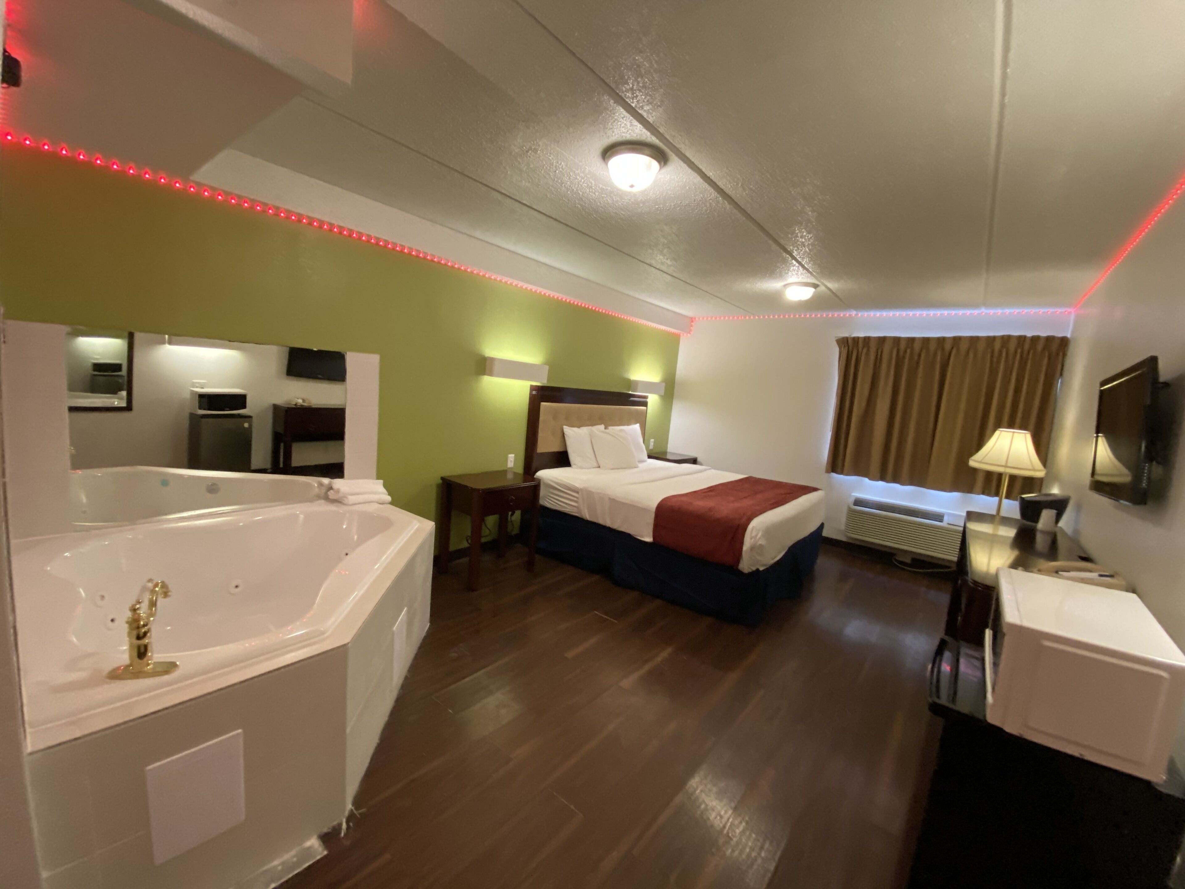 A hotel room with a large bathtub, a bed with a red blanket, a desk with a lamp, and a TV on the wall.