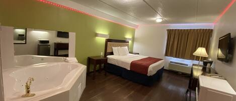 A hotel room with a large bathtub, a bed with a red blanket, a desk with a lamp, and a TV on the wall.