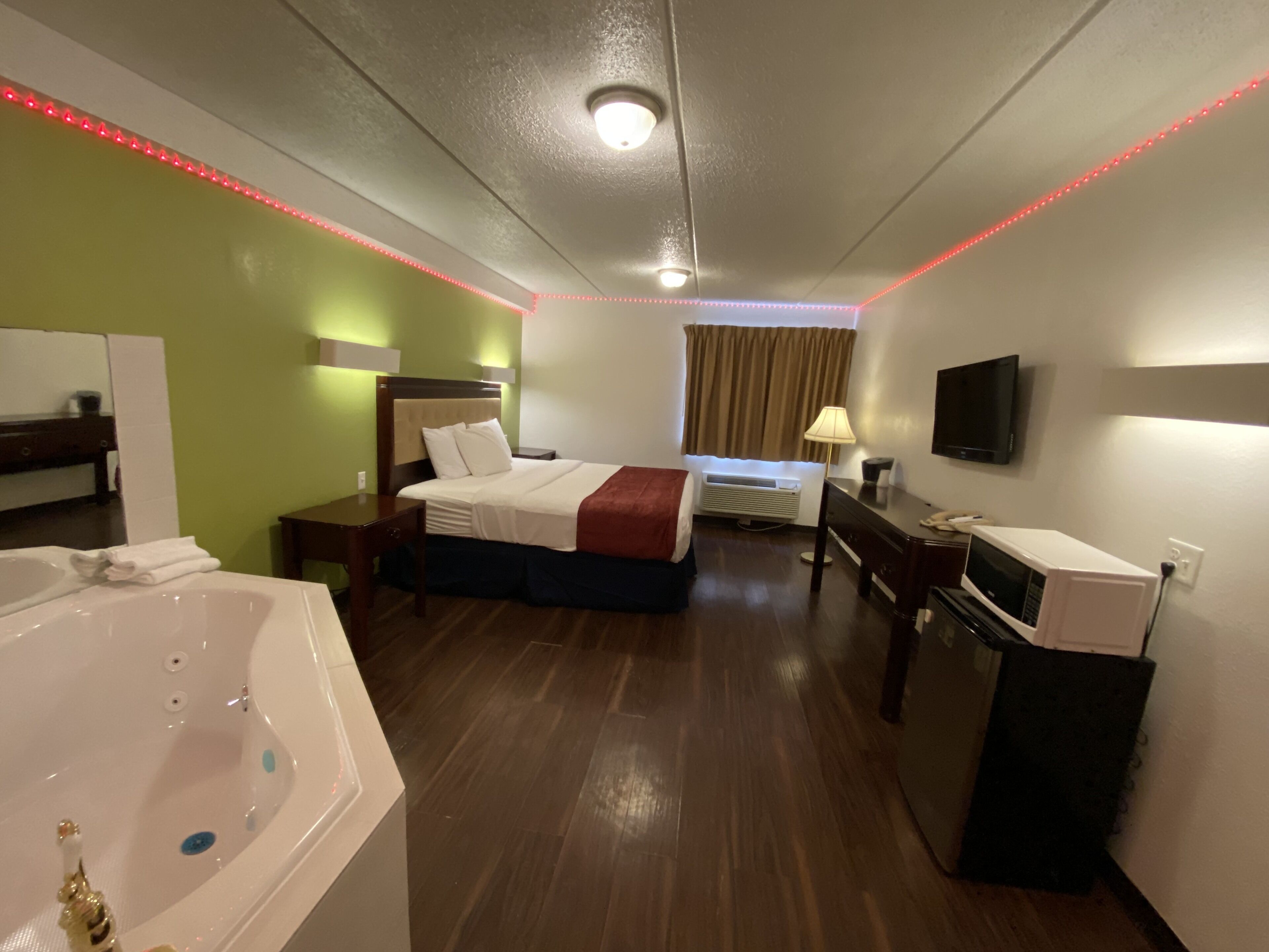 A hotel room with a large bed, a desk, a TV, a microwave, a bathroom with a bathtub, and red LED lights along the ceiling.