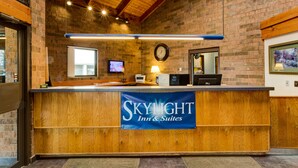 Een receptiebalie met een banner waarop 'SKYLIGHT Inn & Suites' staat, een klok, een televisie en een raam met natuurlijk licht.